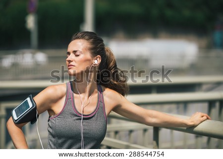 A woman is standing with her eyes closed, basking in the warmth of the sun, and enjoying the moment. Leaning against a guardrail, her head is turned to the side. She is lost in the music.