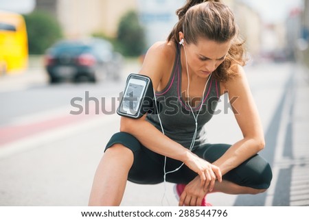 An athletic woman wearing earbuds and her device in a cuff on her arm is looking down at the ground, listening intently to the music.