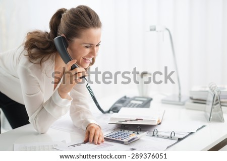 A successful businesswoman is leaning casually on her desk, talking on the telephone, while looking down at a document in a ring-binder.