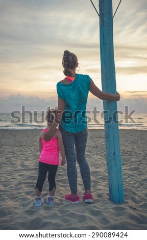 A mother, seen from behind, is standing next to her daughter, resting her hand on her daughter\'s back. They are standing near a flagpole, looking out at the sea at sundown.