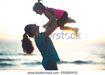 A mother with her hair pulled back in a ponytail is lovingly throwing her child up in the air at sundown. You can practically hear the joyful giggles...