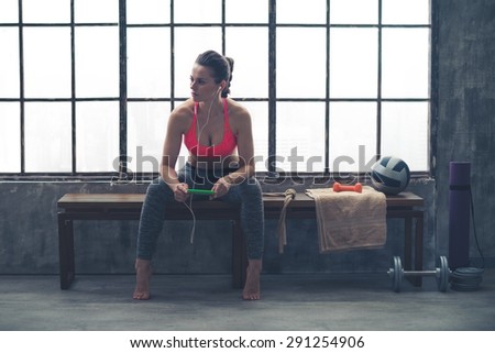 Looking off to the side, an athletic woman watches something in the distance. She is sitting on a bench by a window in a loft gym, holding her device and listening to music.