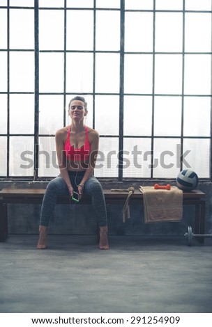 A fit, athletic woman is sitting with her eyes closed, holding her device in her hands. She is lost in the beats and sounds of the amazing music she is listening to as she sits in a loft gym.