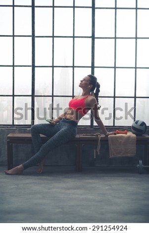 A woman is relaxing on a bench by the window in a big loft gym. Lost in music, she is stretching herself out in a relaxed way, leaning back, head tilted upwards.