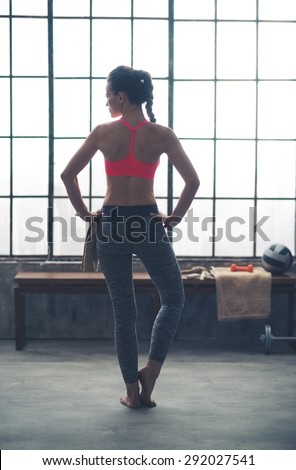 A woman looks off to the side, over her shoulder, at something beyond her in the loft gym. She is standing near a big window and bench.
