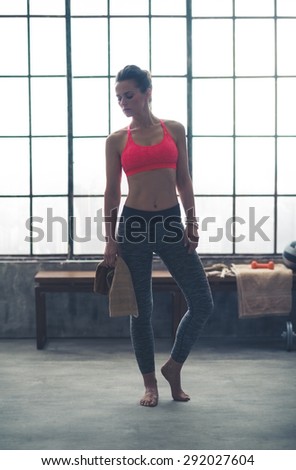 Wearing comfortable workout gear, a woman is standing looking down at the ground in a loft gym, holding a towel. She is barefoot, getting ready to do a yoga workout.