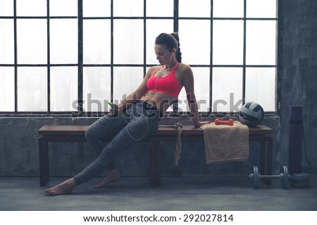 A fit, buff, muscular woman is sitting, relaxed, on a wooden bench in her local loft gym. Looking down at her phone, she is selecting the music she would like to listen to for her workout.