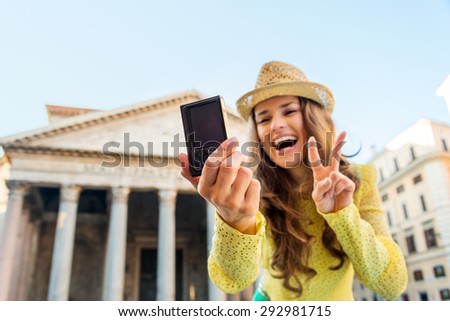 With a focal point on a woman\'s hand holding her digital camera as she is taking a selfie, smiling, and doing the victory sign, a woman tourist takes a photo of herself at the Pantheon in Rome.