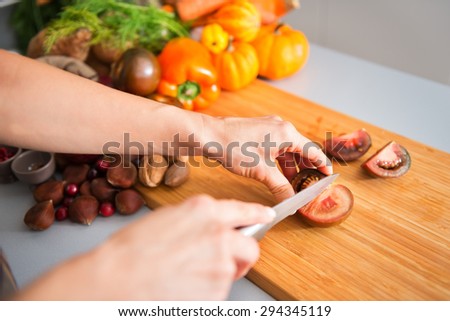 Woman\'s hand slicing a tomato on a wooden cutting board. In the background, a pile of Autumn vegetables, including a bell pepper, miniature pumpkins, chestnuts, cranberries, walnuts, and pecans.