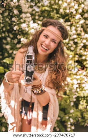 Longhaired hippy-looking young lady in knitted shawl and white blouse standing among flowers with retro camera
