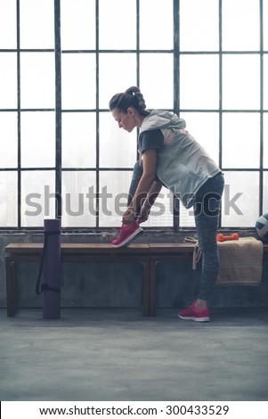 A fit, sporty young woman leans down to tie her shoe, using the bench to help her.