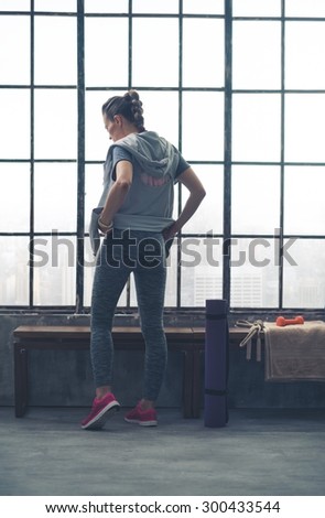 Seen from behind, a fit young woman stands by a city loft gym bench, looking down, adjusting her workout pants.