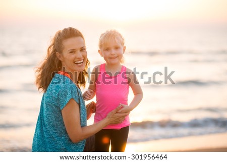 A happy young mother smiles over her shoulder, kneeling next to her young blonde daughter. Both are wearing workout gear and have had fun running together. The sun is going down on a beautiful day.