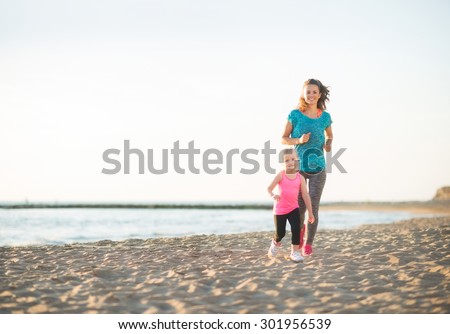 A healthy, fit mother and her young daughter are running together on the beach at sunset. The mother is behind her daughter, who feels like she is winning the race.