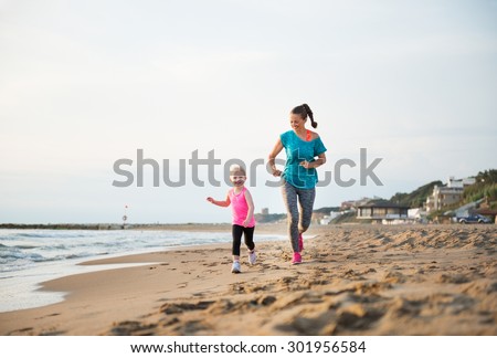 A young girl runs alongside her fit mother, laughing and happy. They are running along the water\'s edge on the beach at sunset. What fun to be out running together!