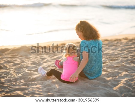 A young mother is sitting next to her daughter, listening to her and playing with her in the sand. Seen from behind, they are both wearing workout gear and are resting, taking time to enjoy the sunset