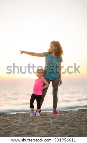 Her young daughter leaning up against her legs, a fit, happy mother points into the distance to show her daughter something. Behind them, the sun is setting on the water.