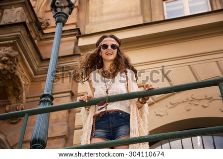 Longhaired hippy-lookin young lady in jeans shorts, knitted shawl and white blouse with sunglasses stands near streetlight in old town looking down and smiling