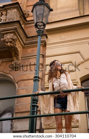 Longhaired hippy-lookin young lady in jeans shorts, knitted shawl and white blouse with sunglasses stands near streetlight in old town looking at skies and smiling