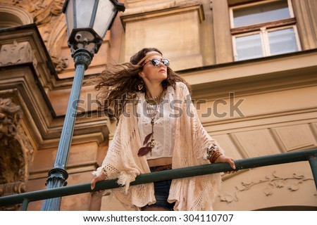 Longhaired hippy young lady in jeans shorts, knitted shawl and white blouse with sunglasses stands near streetlight in old town looking at skies