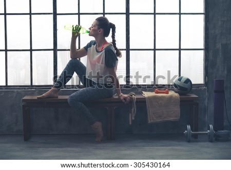 Fit woman in profile sitting on bench in loft gym drinking water. After a good workout, it\'s important to hydrate.