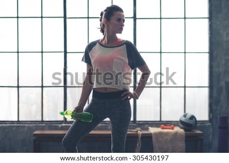 A woman stands relaxing, looking off to the side, holding a water bottle in a loft gym after a good workout.