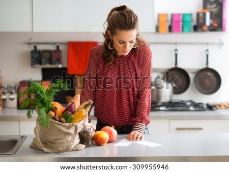 An elegant woman looks down at the shopping lists on her kitchen counter, reading them carefully. Next to her on the kitchen counter, a burlap sac holds a wide variety of fall fruits and vegetables.
