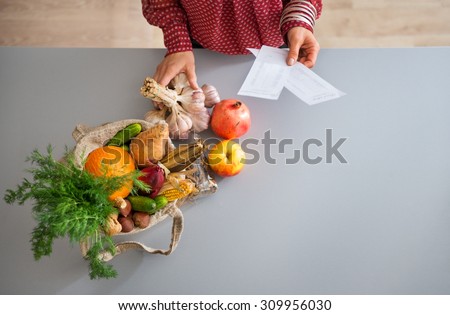 Seen from above, a burlap sac is full of fresh fall fruits and vegetables. A woman\'s elegant hands hold a bunch of fresh garlic and her shopping lists.