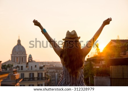 Seen from behind, a woman is standing with outstretched arms, looking out at the city of Rome at sunset in summer.