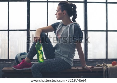 Having had a good workout, a fit, healthy woman takes a few moments to relax and look out at the city below while sitting on a bench holding her water bottle. Next to her, a ball and towel.