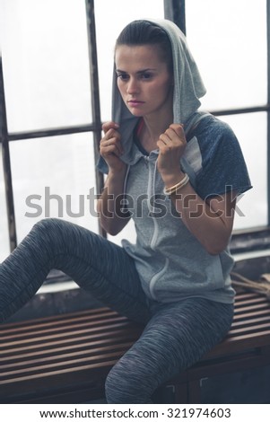 Deep in thought, a woman pauses, sitting on a loft gym bench, while holding her hoodie up on her head, as she thinks about the workout she has just done.