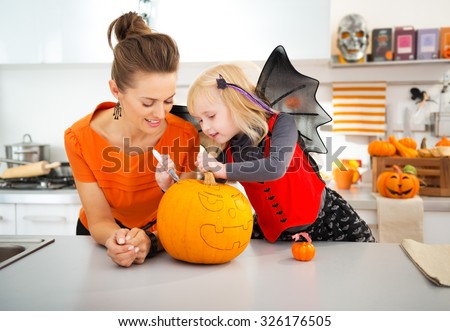 Young mother with daughter in bat costume creating big orange pumpkin Jack-O-Lantern on Halloween party in decorated kitchen. Traditional autumn holiday