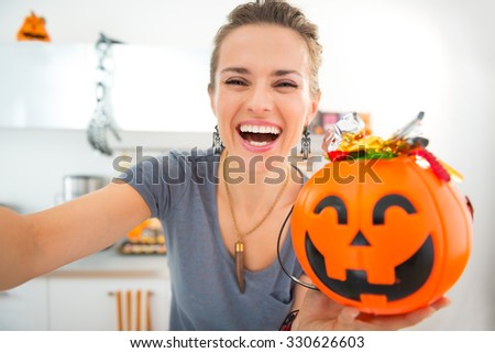 Happy young woman in decorated kitchen making selfie with halloween Jack-o-Lantern bucket full of trick or treat candy. Ready to halloween invasion. Traditional autumn holiday