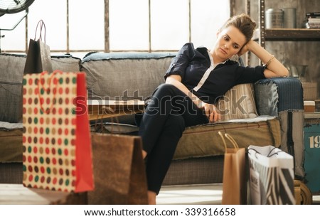 Shopping relieving stress? Tired frustrated brunette woman in elegant clothing sitting on couch among shopping bags in loft apartment.