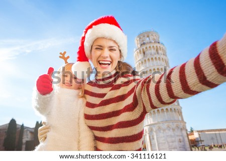 Happy mother in Christmas hat taking selfie with daughter wearing funny reindeer antlers and showing thumbs up in front of Leaning Tour of Pisa, Italy. They spending exciting Christmas time traveling