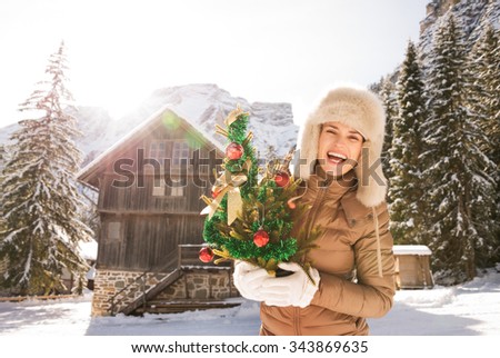 Christmas season in relaxed style of contemporary countryside living. Portrait of smiling young woman holding Christmas tree while standing in the front of a cosy mountain house.