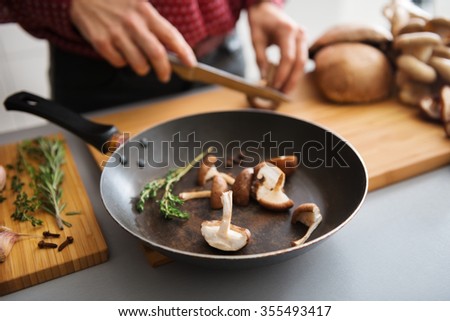 Fresh sliced mushrooms are sitting in a pan on a wooden board, ready to be put on the stove. In the background, a woman slices more fresh mushrooms.