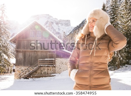Christmas season in relaxed style of contemporary countryside living. Happy young woman standing in the front of a cosy mountain house and looking into the distance