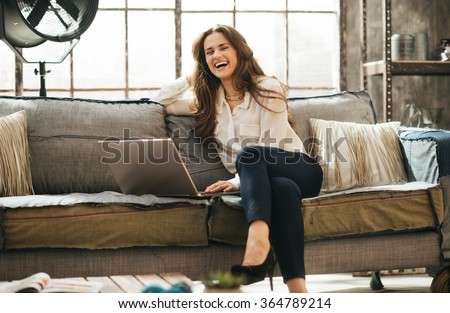 Photo of happy woman in elegant clothing sitting on couch in front open laptop computer in loft living room