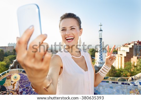 Refreshing promenade in unique Park Guell style in Barcelona, Spain. Portrait of smiling young woman showing victory gesture and taking selfie with smartphone while in Park Guell, Barcelona, Spain