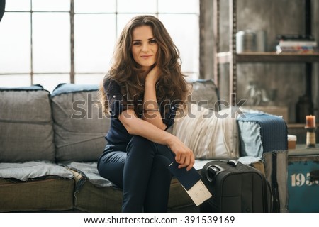 Filling inspired for adventure! Smiling elegant woman with passport, ticket and luggage sitting on sofa in loft apartment while waiting for departure. Happy holiday and travel concept