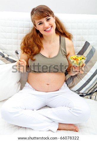 Smiling beautiful pregnant female sitting on sofa with fruit salad