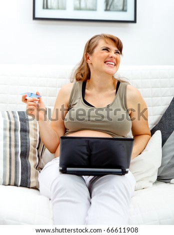 Laughing beautiful pregnant woman sitting on sofa with laptop and credit card