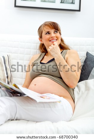 Dreaming beautiful pregnant woman relaxing on sofa at home with magazine.