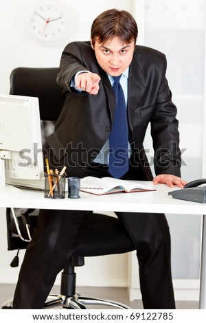 Angry  modern business man standing at office desk and pointing finger on you