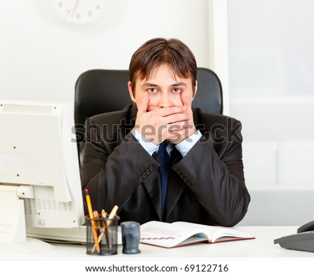 Modern business man sitting at office desk and making speak no evil gesture