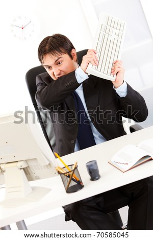 Angry  business man sitting at office desk and destroying computer using keyboard