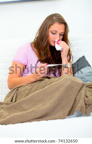 Sick woman with handkerchief in hand sitting on sofa at home and looking at thermometer