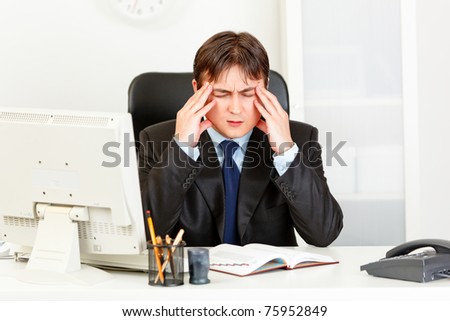 Modern businessman with headache sitting at office desk and holding hands at head