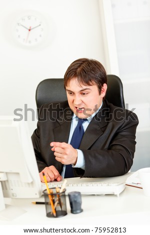 Angry modern businessman sitting at office desk and pointing finger at computer monitor
